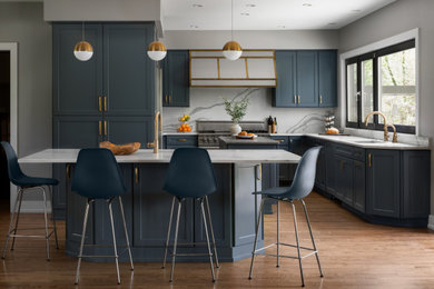 Example of a transitional u-shaped medium tone wood floor and brown floor kitchen design in Milwaukee with an undermount sink, shaker cabinets, blue cabinets, white backsplash, stone slab backsplash, stainless steel appliances, an island and white countertops