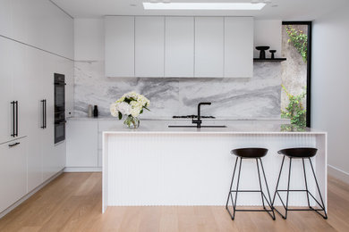 Inspiration for a mid-sized modern galley open plan kitchen in Melbourne with an undermount sink, flat-panel cabinets, white cabinets, solid surface benchtops, grey splashback, marble splashback, black appliances, plywood floors, with island, beige floor, white benchtop and recessed.