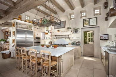 This is an example of a large mediterranean l-shaped open plan kitchen in Orange County with a farmhouse sink, recessed-panel cabinets, distressed cabinets, marble benchtops, white splashback, stainless steel appliances, limestone floors, with island, marble splashback, beige floor and white benchtop.