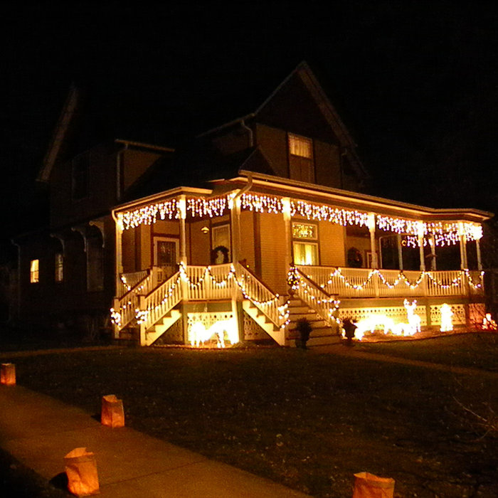 christmas eve night scene with dark sky and queen anne victorian home with wrap-around front porch lit with dangling white lights and wreaths, with paper bag luminaria at the sidewalk
