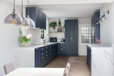 This is an example of a medium sized modern l-shaped kitchen/diner in Surrey with a submerged sink, shaker cabinets, blue cabinets, quartz worktops, white splashback, engineered quartz splashback, black appliances, light hardwood flooring, no island, brown floors, white worktops, exposed beams and feature lighting.