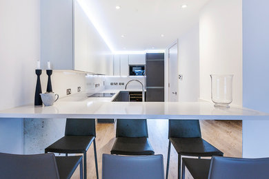 Design ideas for a mid-sized contemporary u-shaped eat-in kitchen in London with a drop-in sink, flat-panel cabinets, white cabinets, quartzite benchtops, white splashback, black appliances, medium hardwood floors, a peninsula and glass sheet splashback.