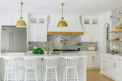 Open concept kitchen - large coastal l-shaped light wood floor and brown floor open concept kitchen idea in Other with a farmhouse sink, shaker cabinets, white cabinets, quartz countertops, gray backsplash, subway tile backsplash, stainless steel appliances, an island and white countertops
