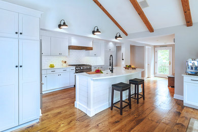 Mid-sized country galley open plan kitchen in Philadelphia with a farmhouse sink, recessed-panel cabinets, white cabinets, quartz benchtops, white splashback, subway tile splashback, stainless steel appliances, medium hardwood floors, with island, brown floor and white benchtop.