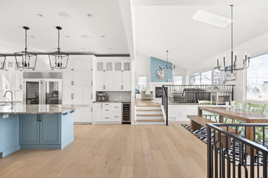 Example of a large trendy u-shaped light wood floor, brown floor and vaulted ceiling eat-in kitchen design in Calgary with an undermount sink, raised-panel cabinets, white cabinets, granite countertops, white backsplash, ceramic backsplash, stainless steel appliances, an island and multicolored countertops