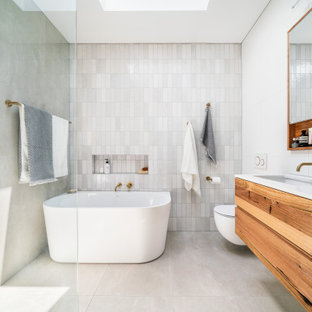 This is an example of a small contemporary master bathroom in Melbourne with furniture-like cabinets, medium wood cabinets, a freestanding tub, an open shower, a wall-mount toilet, gray tile, mosaic tile, white walls, porcelain floors, an undermount sink, solid surface benchtops, grey floor, an open shower, white benchtops, a shower seat, a single vanity and a floating vanity.