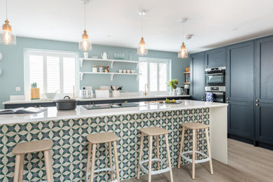 Large nautical l-shaped open plan kitchen in Cornwall with a built-in sink, shaker cabinets, blue cabinets, engineered stone countertops, white splashback, engineered quartz splashback, black appliances, vinyl flooring, an island, grey floors, white worktops and a feature wall.