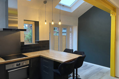 Design ideas for a small contemporary open plan kitchen in Manchester with flat-panel cabinets, black cabinets, laminate countertops, black splashback, stainless steel appliances, laminate floors, an island, beige floors, exposed beams and feature lighting.