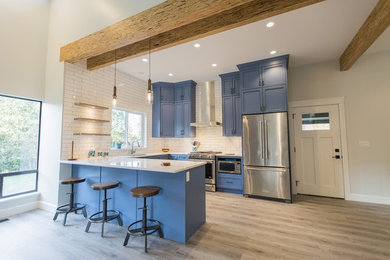This is an example of a mid-sized country u-shaped kitchen in Vancouver with an undermount sink, recessed-panel cabinets, blue cabinets, quartzite benchtops, white splashback, subway tile splashback, stainless steel appliances, light hardwood floors, a peninsula, beige floor and white benchtop.