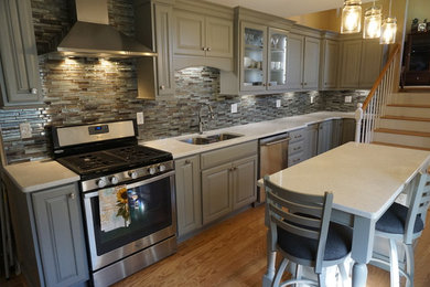 Photo of a small traditional l-shaped eat-in kitchen in Providence with a triple-bowl sink, raised-panel cabinets, grey cabinets, quartz benchtops, grey splashback, mosaic tile splashback, stainless steel appliances, medium hardwood floors, with island and white benchtop.