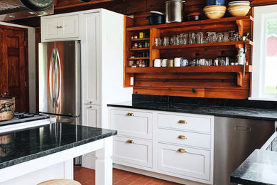 This is an example of a mid-sized country l-shaped kitchen in New York with a farmhouse sink, beaded inset cabinets, white cabinets, soapstone benchtops, stainless steel appliances, ceramic floors, with island, red floor and black benchtop.