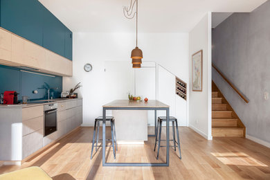 Mid-sized contemporary galley open plan kitchen in Paris with a single-bowl sink, beaded inset cabinets, light wood cabinets, concrete benchtops, green splashback, glass sheet splashback, panelled appliances, light hardwood floors, with island and grey benchtop.