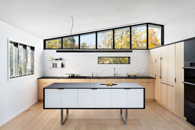 Trendy light wood floor and vaulted ceiling kitchen photo in New York with an undermount sink, flat-panel cabinets, light wood cabinets, laminate countertops, white backsplash, ceramic backsplash, black appliances, an island and black countertops