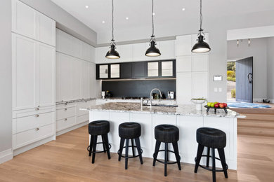 Large farmhouse l-shaped open plan kitchen in Auckland with a double-bowl sink, shaker cabinets, white cabinets, granite worktops, black splashback, integrated appliances, light hardwood flooring and an island.