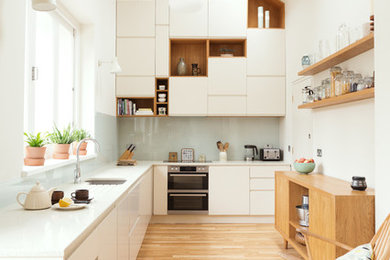 Design ideas for a mid-sized contemporary l-shaped kitchen in London with flat-panel cabinets, white cabinets, recycled glass benchtops, blue splashback, glass sheet splashback, no island, white benchtop, an undermount sink, light hardwood floors and beige floor.