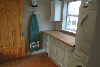 This is an example of a classic single-wall utility room in Cambridgeshire with a single-bowl sink, raised-panel cabinets, white cabinets, wood worktops, beige walls, terracotta flooring and a stacked washer and dryer.