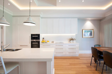 Photo of a mid-sized transitional galley open plan kitchen in Sydney with an undermount sink, shaker cabinets, white cabinets, solid surface benchtops, white splashback, subway tile splashback, black appliances, medium hardwood floors, a peninsula, brown floor and white benchtop.