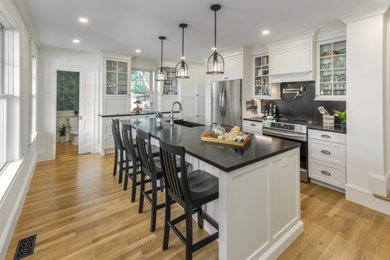 Inspiration for a large timeless l-shaped medium tone wood floor and beige floor enclosed kitchen remodel in Providence with a farmhouse sink, recessed-panel cabinets, white cabinets, wood countertops, black backsplash, stone slab backsplash, stainless steel appliances, an island and black countertops