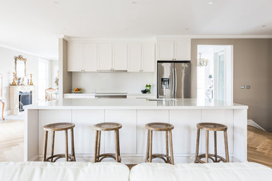 Large contemporary single-wall open plan kitchen in Melbourne with an undermount sink, shaker cabinets, white cabinets, quartz benchtops, white splashback, stone slab splashback, stainless steel appliances, medium hardwood floors, with island, brown floor and white benchtop.