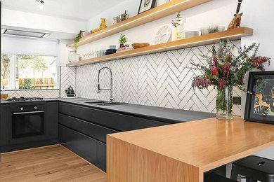 This is an example of a mid-sized modern u-shaped eat-in kitchen in Melbourne with a double-bowl sink, flat-panel cabinets, black cabinets, solid surface benchtops, white splashback, porcelain splashback, black appliances, light hardwood floors, with island and black benchtop.