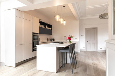 Mid-sized contemporary l-shaped open plan kitchen in London with an undermount sink, flat-panel cabinets, beige cabinets, quartzite benchtops, white splashback, engineered quartz splashback, stainless steel appliances, light hardwood floors, with island, grey floor and white benchtop.