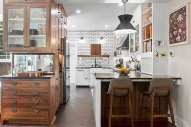 Example of a mid-sized mid-century modern slate floor and black floor kitchen design in Philadelphia with a farmhouse sink, glass-front cabinets, dark wood cabinets, white backsplash, subway tile backsplash, stainless steel appliances, a peninsula, black countertops and soapstone countertops
