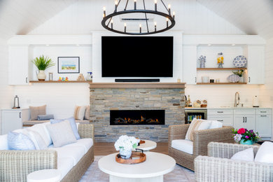 Example of a beach style medium tone wood floor, brown floor, vaulted ceiling and shiplap wall living room design in New York with white walls, a ribbon fireplace, a stacked stone fireplace and a wall-mounted tv