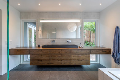 Photo of a large contemporary master bathroom in Los Angeles with furniture-like cabinets, medium wood cabinets, a drop-in tub, a wall-mount toilet, white tile, ceramic tile, white walls, a vessel sink and wood benchtops.