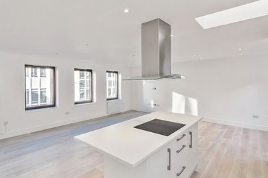 Photo of a modern kitchen in London with white cabinets, solid surface benchtops, grey splashback, glass sheet splashback, stainless steel appliances, with island, white benchtop, laminate floors and beige floor.
