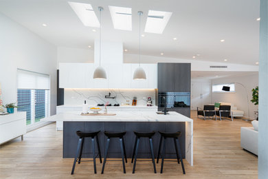 Photo of a large modern eat-in kitchen in Sydney with a single-bowl sink, flat-panel cabinets, white cabinets, quartz benchtops, white splashback, engineered quartz splashback, black appliances, vinyl floors, with island, brown floor, white benchtop and vaulted.