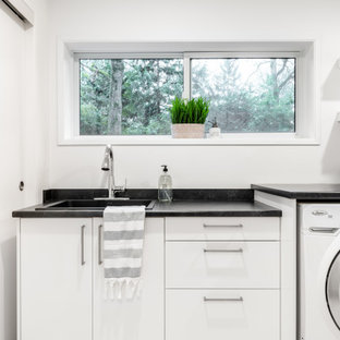 バンクーバーの中サイズのミッドセンチュリースタイルのおしゃれな洗濯室 (ドロップインシンク、フラットパネル扉のキャビネット、白いキャビネット、ラミネートカウンター、白い壁、クッションフロア、左右配置の洗濯機・乾燥機、グレーの床、黒いキッチンカウンター) の写真