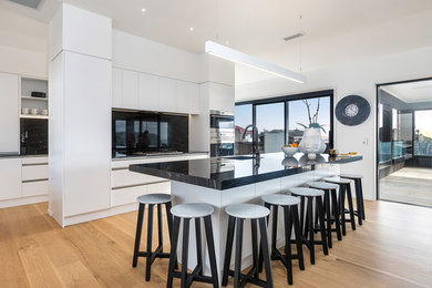 Photo of a large modern galley open plan kitchen in Auckland with an undermount sink, flat-panel cabinets, white cabinets, granite benchtops, black splashback, stone slab splashback, stainless steel appliances, light hardwood floors, with island, beige floor and black benchtop.