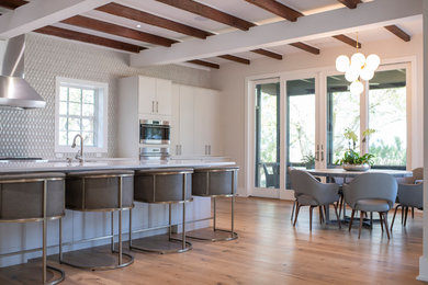 Inspiration for a large contemporary l-shaped medium tone wood floor, brown floor, exposed beam and vaulted ceiling eat-in kitchen remodel in Charleston with flat-panel cabinets, white cabinets, stainless steel appliances, an island, white countertops, gray backsplash, quartzite countertops, porcelain backsplash and a farmhouse sink