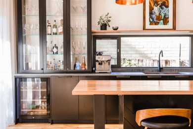 Inspiration for an industrial galley kitchen pantry in Sydney with a farmhouse sink, flat-panel cabinets, black cabinets, wood benchtops, grey splashback, engineered quartz splashback, black appliances, light hardwood floors and multiple islands.