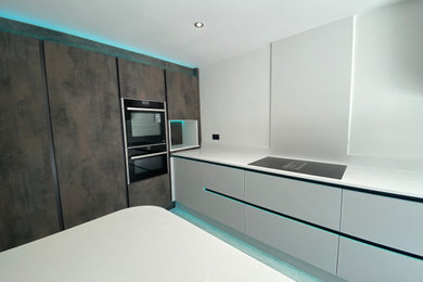 This is an example of a large modern grey and black l-shaped enclosed kitchen in Other with an integrated sink, flat-panel cabinets, grey cabinets, composite countertops, white splashback, engineered quartz splashback, black appliances, light hardwood flooring, a breakfast bar, grey floors, white worktops and feature lighting.