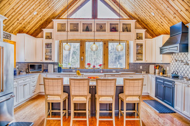 Example of a large beach style u-shaped medium tone wood floor, brown floor, vaulted ceiling and wood ceiling eat-in kitchen design in New York with a farmhouse sink, recessed-panel cabinets, white cabinets, quartz countertops, blue backsplash, porcelain backsplash, an island and white countertops