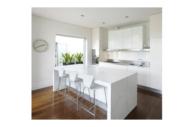 Photo of a mid-sized modern single-wall eat-in kitchen in Sydney with a double-bowl sink, recessed-panel cabinets, white cabinets, stainless steel benchtops, white splashback, ceramic splashback, stainless steel appliances, medium hardwood floors and with island.