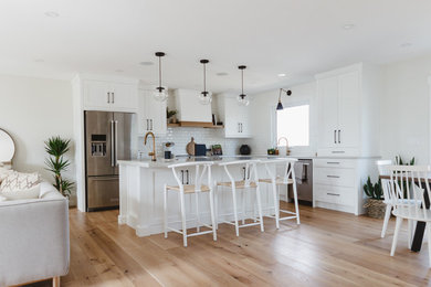 Mid-sized transitional l-shaped eat-in kitchen in Other with an undermount sink, shaker cabinets, white cabinets, quartz benchtops, white splashback, ceramic splashback, stainless steel appliances, light hardwood floors, with island, brown floor and white benchtop.