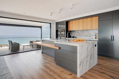 Design ideas for a large modern single-wall open plan kitchen in Sydney with an undermount sink, flat-panel cabinets, grey cabinets, quartzite benchtops, grey splashback, stone slab splashback, black appliances, medium hardwood floors, with island, brown floor and grey benchtop.