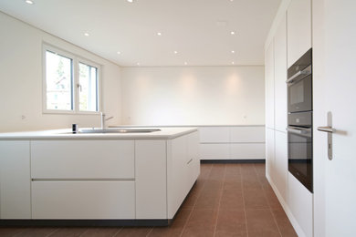 Offene, Große Moderne Küche mit Unterbauwaschbecken, flächenbündigen Schrankfronten, weißen Schränken, Mineralwerkstoff-Arbeitsplatte, Küchenrückwand in Weiß, schwarzen Elektrogeräten, Keramikboden, Kücheninsel, braunem Boden, weißer Arbeitsplatte und eingelassener Decke in Sonstige