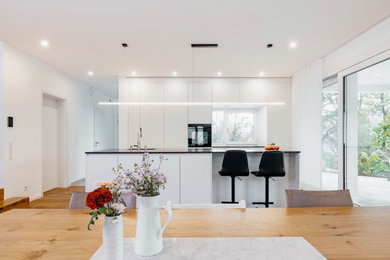 Zweizeilige, Mittelgroße Moderne Küche mit Waschbecken, flächenbündigen Schrankfronten, weißen Schränken, Granit-Arbeitsplatte, schwarzen Elektrogeräten, braunem Holzboden, Kücheninsel, braunem Boden, grauer Arbeitsplatte und Tapetendecke in Frankfurt am Main