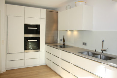 Moderne Küche in L-Form mit weißen Schränken, Mineralwerkstoff-Arbeitsplatte, Küchenrückwand in Weiß und Glasrückwand in München