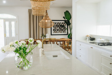 Inspiration for a mid-sized traditional galley open plan kitchen in Brisbane with a double-bowl sink, recessed-panel cabinets, white cabinets, quartz benchtops, white splashback, black appliances, medium hardwood floors, with island, brown floor and beige benchtop.