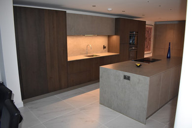 Large contemporary single-wall open plan kitchen in Kent with an integrated sink, flat-panel cabinets, dark wood cabinets, concrete worktops, grey splashback, stone tiled splashback, black appliances, porcelain flooring, an island, white floors and grey worktops.