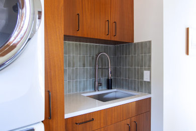 Inspiration for a mid-sized slate floor and black floor laundry room remodel in Minneapolis with a drop-in sink, flat-panel cabinets, medium tone wood cabinets, granite countertops, blue backsplash, subway tile backsplash, white walls, a stacked washer/dryer and white countertops