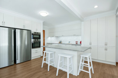 Inspiration for a mid-sized beach style galley eat-in kitchen in Newcastle - Maitland with an undermount sink, recessed-panel cabinets, white cabinets, laminate benchtops, white splashback, ceramic splashback, stainless steel appliances, laminate floors, with island and brown floor.