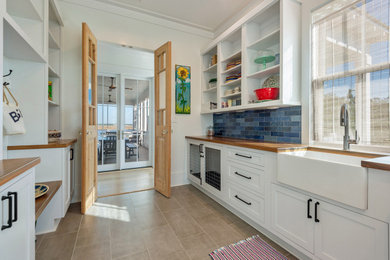Inspiration for a mid-sized transitional l-shaped ceramic tile and beige floor utility room remodel in New Orleans with a farmhouse sink, shaker cabinets, white cabinets, wood countertops, blue backsplash, glass tile backsplash, white walls and brown countertops