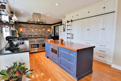 Example of a large farmhouse u-shaped medium tone wood floor and brown floor eat-in kitchen design in New York with a farmhouse sink, recessed-panel cabinets, white cabinets, wood countertops, stainless steel appliances, an island, brown countertops, white backsplash and subway tile backsplash