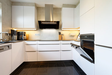 This is an example of a small contemporary u-shaped separate kitchen in Essen with a drop-in sink, flat-panel cabinets, white cabinets, white splashback, glass sheet splashback, black appliances, no island and black floor.