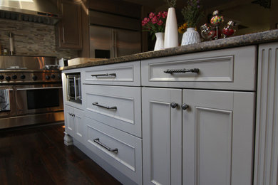 Large trendy l-shaped dark wood floor eat-in kitchen photo in New York with a drop-in sink, raised-panel cabinets, gray cabinets, granite countertops, gray backsplash, stone tile backsplash, stainless steel appliances and an island